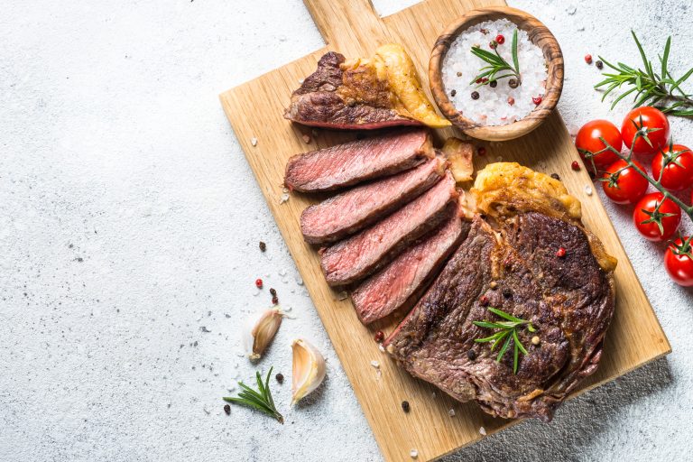 Grilled beef steak ribeye medium rare on wooden cutting board on white stone table. Top view with copy space.